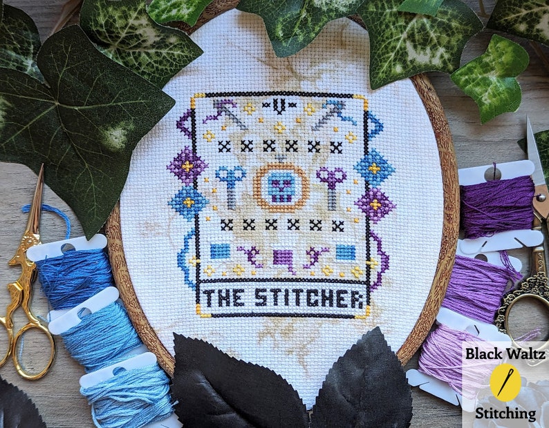 May include: A white embroidery hoop with a cross-stitch design featuring a skull, keys, and scissors. The design is surrounded by a colorful border and the text "The Stitcher" is below the design.