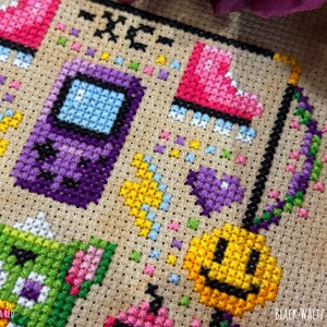 May include: Cross-stitch embroidery featuring a purple handheld game console, pink roller skate, green creature, and yellow smiley face. The design includes colorful accents and the text "-XC-" and "TEST STITCH - SARA RED". The words "BLACK WALTZ STITCHING" are also visible.