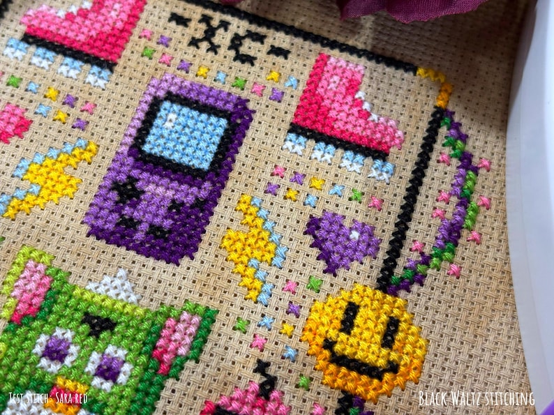 May include: Cross-stitch embroidery featuring a purple handheld game console, pink roller skate, green creature, and yellow smiley face. The design includes colorful accents and the text "-XC-" and "TEST STITCH - SARA RED". The words "BLACK WALTZ STITCHING" are also visible.