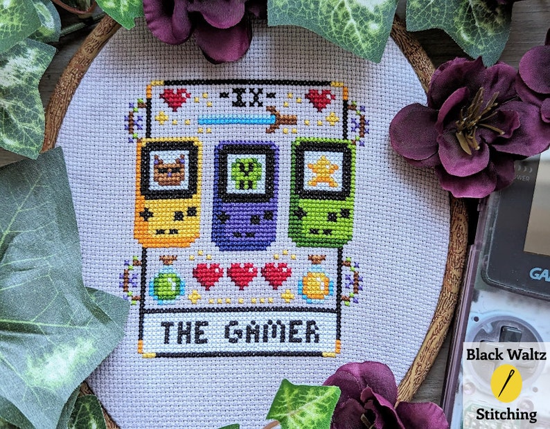 May include: A cross-stitch embroidery hoop with a white background and a colorful pixelated design of three handheld video game consoles. The design includes the text "THE GAMER".