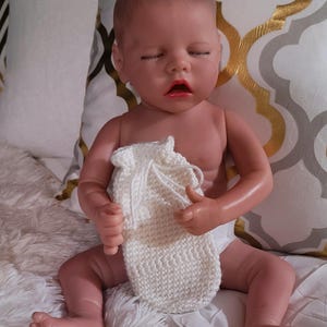 May include: A white knitted baby bootie with a drawstring closure. The bootie is being held by a baby doll with closed eyes and a pink mouth.