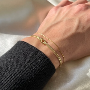Op de afbeelding: Twee gouden armbanden om een pols. De ene armband is een dunne ketting, de andere is een dikkere ketting met een knoopdesign.