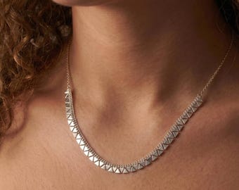 May include: A silver-toned necklace featuring a series of small, triangular pendants. The necklace is delicate and sits close to the neck, creating a minimalist and modern look. The chain is fine, and the triangles are evenly spaced.