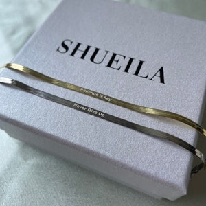 May include: Two gold and silver chain necklaces with engraved messages. The gold chain reads "Patience is key" and the silver chain reads "Never Give Up". The necklaces are displayed on a white box with the brand name "SHUEILA" printed on the top.