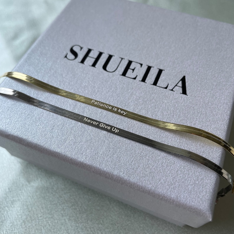 May include: Two gold and silver chain necklaces with engraved messages. The gold chain reads "Patience is key" and the silver chain reads "Never Give Up". The necklaces are displayed on a white box with the brand name "SHUEILA" printed on the top.