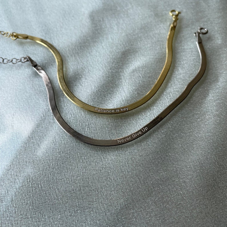 May include: Two gold and silver snake chain necklaces with engraved messages. The gold necklace says "Patience is key" and the silver necklace says "Never Give Up".