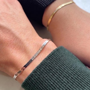Puede incluir: Se muestran dos pulseras, una plateada y otra dorada. La pulsera plateada tiene grabadas las coordenadas "41.0137009 N". La pulsera dorada tiene texto grabado. Ambas pulseras son finas y se llevan en la muñeca.