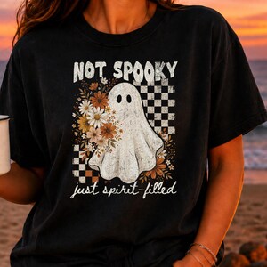 May include: A black t-shirt featuring a graphic of a white ghost surrounded by orange and white flowers, a black and white checkered pattern, and the text "NOT SPOOKY" at the top and "just spirit-filled" at the bottom in a white script font.