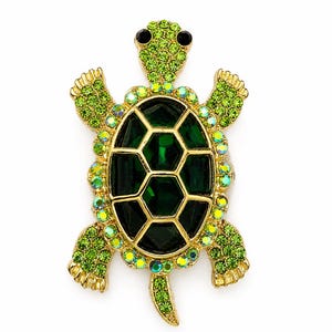 May include: A gold-toned turtle brooch featuring a central shell made of dark green faceted stones. The head, limbs, and tail are encrusted with small lime green crystals, while the border of the shell is lined with iridescent, multi-colored rhinestones.