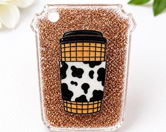 Può includere: Un portachiavi in acrilico trasparente a forma di tazza da caffè riempito di glitter oro rosa. Al centro dei glitter si trova la grafica di una tazza da caffè con coperchio nero, corpo a griglia color cuoio e fascia bianca con macchie di mucca.