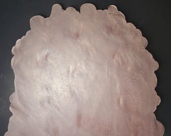 Può includere: Un sottobicchiere rosa chiaro di forma irregolare con una superficie strutturata. Il sottobicchiere ha un bordo smerlato ed è posto su uno sfondo scuro. Il sottobicchiere sembra essere fatto di resina.