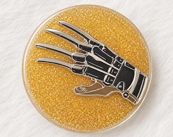 Puede incluir: Un pin circular con un fondo de purpurina dorada y un diseño central de guante metálico negro y plateado con cuatro hojas largas y afiladas que se extienden desde los dedos. El accesorio tiene un acabado liso, brillante y un borde claro.