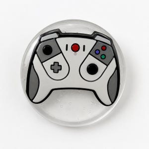 May include: A circular pin featuring a white and grey video game controller graphic. The controller design includes a central red button, a black directional pad, a black joystick, and three smaller coloured buttons in red, blue, and green.