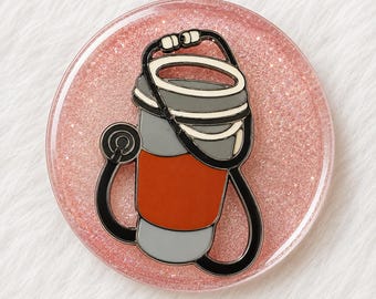 Può includere: Una spilla circolare in resina glitterata rosa con un design centrale in smalto raffigurante una tazza da caffè grigia e arancione avvolta da uno stetoscopio nero. La tazza ha un coperchio bianco e una fascia arancione nella parte centrale.