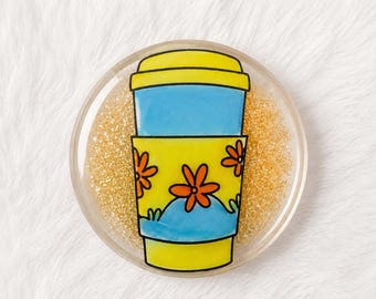 Può includere: Un sottobicchiere circolare con una base in resina trasparente riempita di glitter dorati. Al centro, è raffigurata un'illustrazione grafica di un bicchiere da caffè blu e giallo, decorato con motivi floreali arancioni su una fascia gialla.