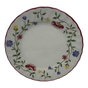 May include: A round white ceramic plate with a scalloped edge and a thin dark pink rim. The plate features a delicate floral pattern with green vines, small blue bell-shaped flowers, yellow blossoms, and larger red and pink flowers.