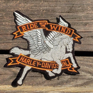 Peut inclure: Écusson brodé avec un cheval ailé, le texte "RIDE WITH THE WIND" et "HARLEY-DAVIDSON" en orange et blanc. L'écusson a une bordure noire. Sur fond de bois.