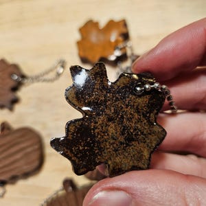 May include: Close-up of several leaf-shaped keychains. The keychains are made of a dark brown material with a speckled, textured appearance. Each keychain has a silver-colored chain attached. The leaves have a glossy finish and are held in a hand.
