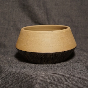May include: A brown ceramic planter with a dark brown, textured base. The planter is sitting on a gray fabric surface.