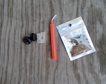 May include: An orange candle, two black stones, a clear crystal, and a small bag of herbs are arranged on a weathered wooden surface. The items are likely used for spiritual or ritualistic purposes.