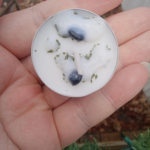 May include: A small, round, white candle in a silver metal container. The candle has a textured surface with two dark blue stones and green herbs. A small, white wick is visible in the centre.