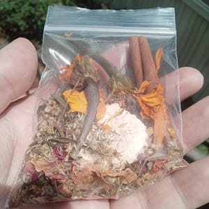Może przedstawiać: Przezroczysta plastikowa torba wypełniona suszonymi ziołami, kwiatami i laskami cynamonu. Zawartość obejmuje pomarańczowe i żółte płatki, brązowe laski cynamonu oraz różne zielone i brązowe suszone zioła. Torba jest trzymana w dłoni.