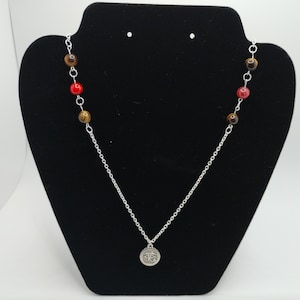 May include: A silver chain necklace with a silver pendant featuring an eye symbol. The necklace is adorned with brown and red beads.
