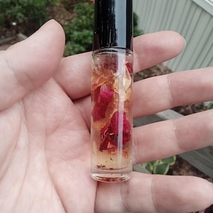 Puede incluir: Un frasco de rodillo de vidrio transparente con una tapa negra, lleno de un líquido de color claro y pétalos de rosa rojos. El frasco se sostiene en una mano, con un fondo borroso de vegetación.