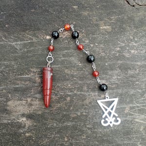 May include: A rosary-style necklace featuring a red, pointed pendant and a chain of black and red beads. The necklace has a silver-colored charm with a stylized symbol at the end. The beads are strung on a silver-colored chain.