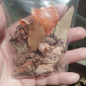 Può includere: Un sacchetto di plastica trasparente pieno di erbe e spezie essiccate. Il contenuto comprende scorza d'arancia, foglie di alloro e altre materie vegetali essiccate. Il sacchetto è tenuto in mano, con una superficie di legno sullo sfondo.