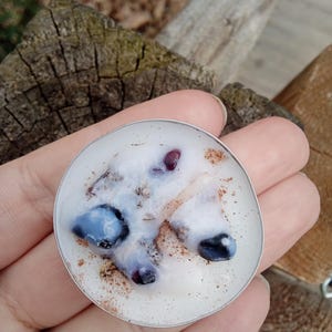 May include: A circular pendant with a silver border, featuring a white resin interior. It contains blue and purple stones, small red details, and brown flecks. The pendant is held in a hand.