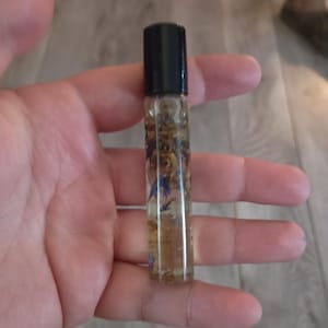 Peut inclure: Un flacon à bille en verre transparent avec un bouchon noir, rempli d'un liquide contenant de petits éléments botaniques séchés, dont des pétales de fleurs bleus et jaunes en suspension dans le mélange huileux transparent.