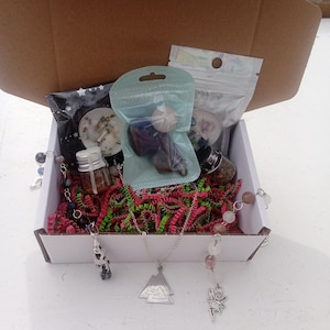 May include: An open cardboard box filled with various items, including small bags, bottles, and necklaces. The box contains red and green shredded paper. The necklaces have silver pendants. The items are arranged inside a white box.