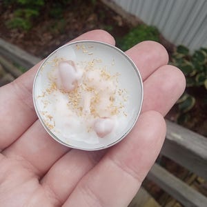 Puede incluir: Una pequeña vela redonda en un recipiente de metal plateado. La cera de la vela es blanca con decoraciones en forma de corazón rosa y pequeñas chispas de color marrón claro. La vela se sostiene en una mano, con un fondo borroso al aire libre.