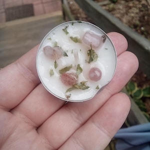 Puede incluir: Una pequeña vela redonda en un recipiente de metal plateado. La cera blanca está decorada con pequeñas piedras rosas y hierbas verdes. La vela se sostiene en una mano abierta, con un fondo borroso al aire libre.