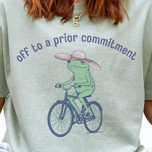 Puede incluir: Camiseta verde salvia con una rana de dibujos animados en bicicleta, con sombrero. El texto "off to a prior commitment" está encima de la rana. La rana es verde, la bicicleta es azul y el sombrero es rosa.