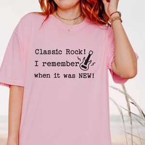 May include: A light pink short-sleeved t-shirt featuring black text that reads "Classic Rock! I remember when it was NEW!" accompanied by a small black graphic of an electric guitar with lightning bolt accents on the front of the garment.