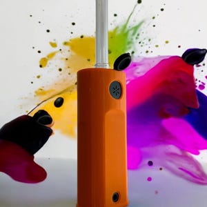 May include: An orange and clear glass vaporizer with a black mouthpiece and a black button. The vaporizer is on a white surface with a colorful background.