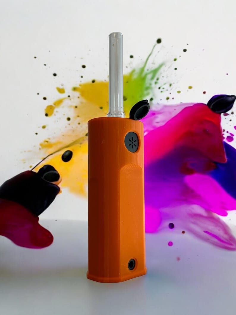 May include: An orange and clear glass vaporizer with a black mouthpiece and a black button. The vaporizer is on a white surface with a colorful background.