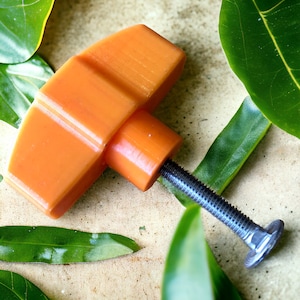 May include: Orange plastic knob with a threaded metal screw. The knob is on a light brown surface with green leaves.