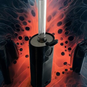 May include: A black and silver vaporizer with a glass mouthpiece. The vaporizer is sitting on a red and black background.