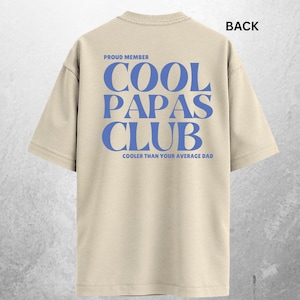 May include: Ivory-colored t-shirt with the text "COOL PAPAS CLUB" in blue block letters on the back. The text "PROUD MEMBER" is above the main text, and "COOLER THAN YOUR AVERAGE DAD" is below. The shirt is a casual style.