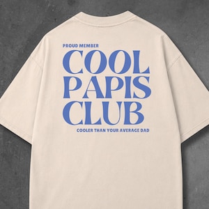 May include: A beige short-sleeve t-shirt featuring blue text on the back. The text reads PROUD MEMBER COOL PAPIS CLUB COOLER THAN YOUR AVERAGE DAD. The shirt has a crew neckline and a standard fit, displaying the back side of the garment clearly.