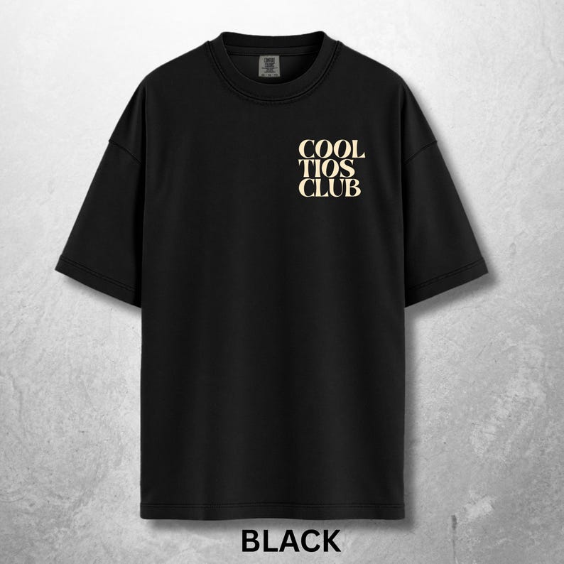 May include: Black t-shirt with the text "COOL TIOS CLUB" in a light tan color on the upper chest. The word "BLACK" is printed in white at the bottom of the shirt. The shirt has short sleeves and a crew neck.