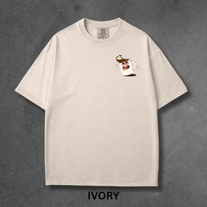 May include: Ivory-colored t-shirt with a small cartoon coffee cup graphic on the chest. The shirt is a relaxed fit with short sleeves and a crew neck. The word "IVORY" is printed at the bottom of the image.