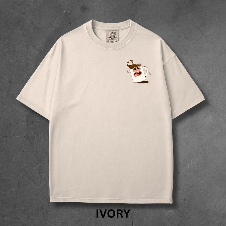 May include: Ivory-colored t-shirt with a small cartoon coffee cup graphic on the chest. The shirt is a relaxed fit with short sleeves and a crew neck. The word "IVORY" is printed at the bottom of the image.