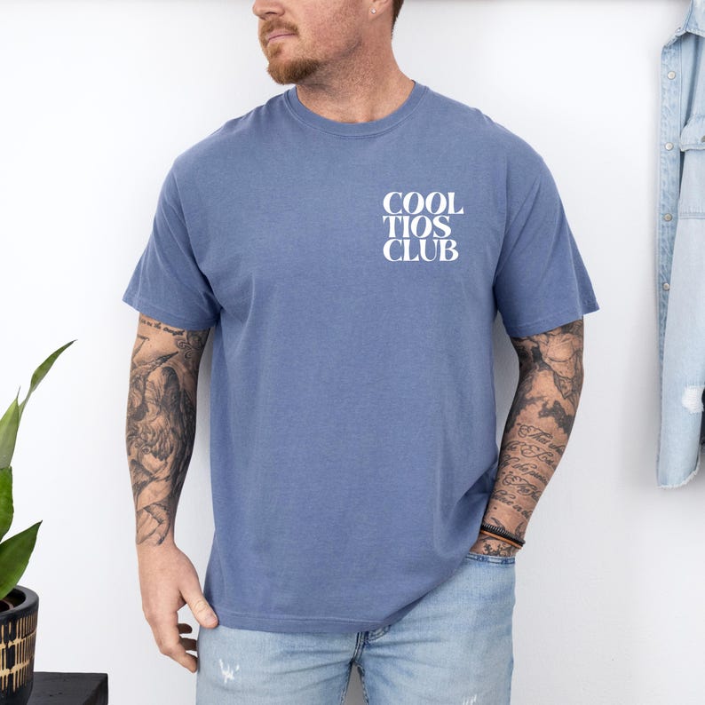 May include: A blue short-sleeve t-shirt with the text "COOL TIOS CLUB" in white block letters on the upper left chest. The shirt is a solid color and appears to be made of a soft material.