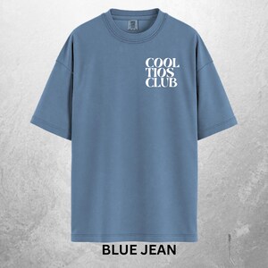 May include: A blue jean colored t-shirt with the text "COOL TIOS CLUB" in white on the front. The shirt has a crew neck and short sleeves. The words "BLUE JEAN" are printed in white at the bottom of the shirt.