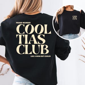 May include: Black sweatshirt with the text "COOL TIAS CLUB" in a cream-colored font. The text "PROUD MEMBER" is above the main text, and "LIKE A MOM BUT COOLER" is below. A smaller version of the text is on the sleeve.