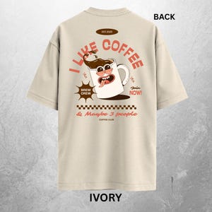 May include: Ivory-colored t-shirt with a coffee-themed graphic on the back. The design features a cartoon coffee mug with the text "I Like Coffee" and "Brew Crew." Additional text includes "& Maybe 3 people" and "Join Now!"